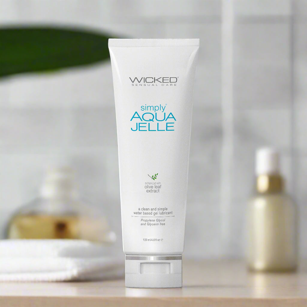 A white tube of Wicked Sensual Care Simply Aqua Jelle water-based lubricant stands upright on a bathroom counter, with blurred toiletries and a green leaf in the background.