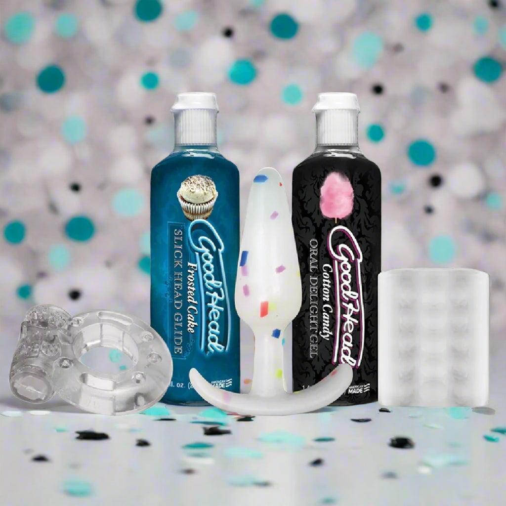 Two bottles of GoodHead gel, a white dildo, and two clear textured sleeves are arranged upright on a surface with blue, black, and white confetti in the background.