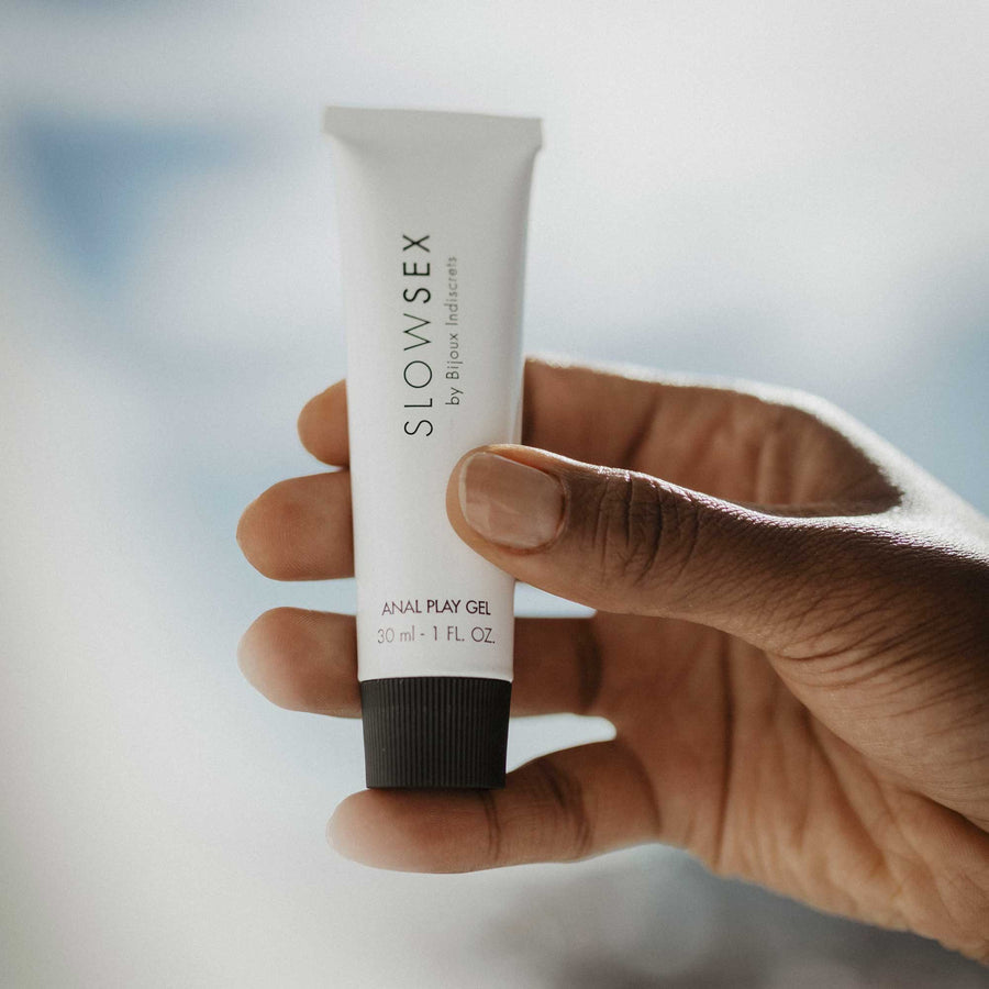 A hand holds a small white tube labeled "SLOW SEX by Bijoux Indiscrets, Anal Play Gel, 30 ml - 1 fl oz," against a softly blurred background.