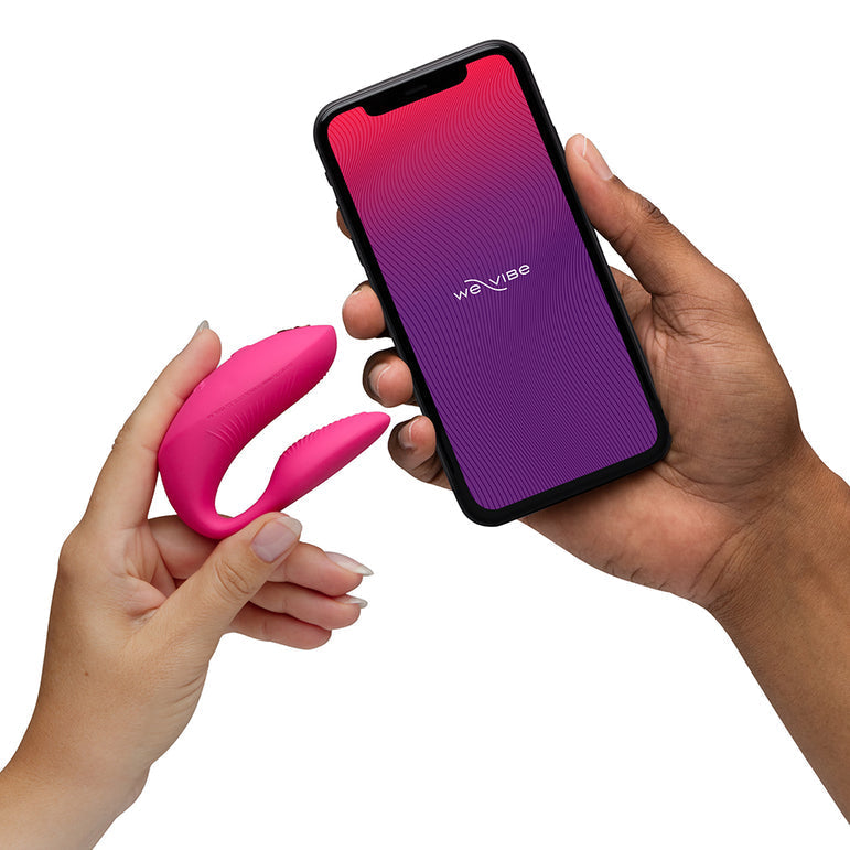 A person holds a pink wearable vibrator, while another person holds a smartphone displaying the We-Vibe app on the screen.