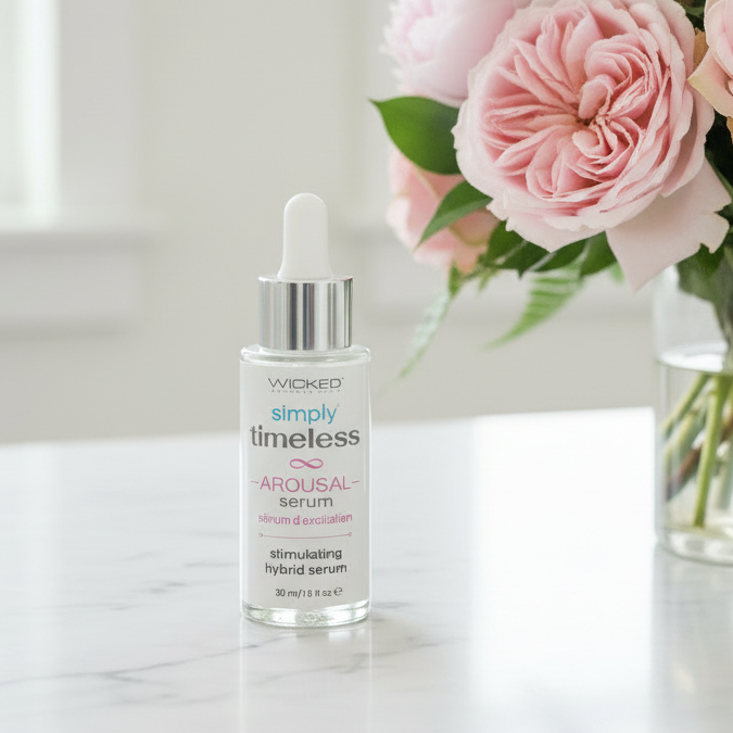 A dropper bottle of WIOKED Simply Timeless Arousal Serum sits on a white surface beside a glass vase of pink roses, with a soft, bright background.