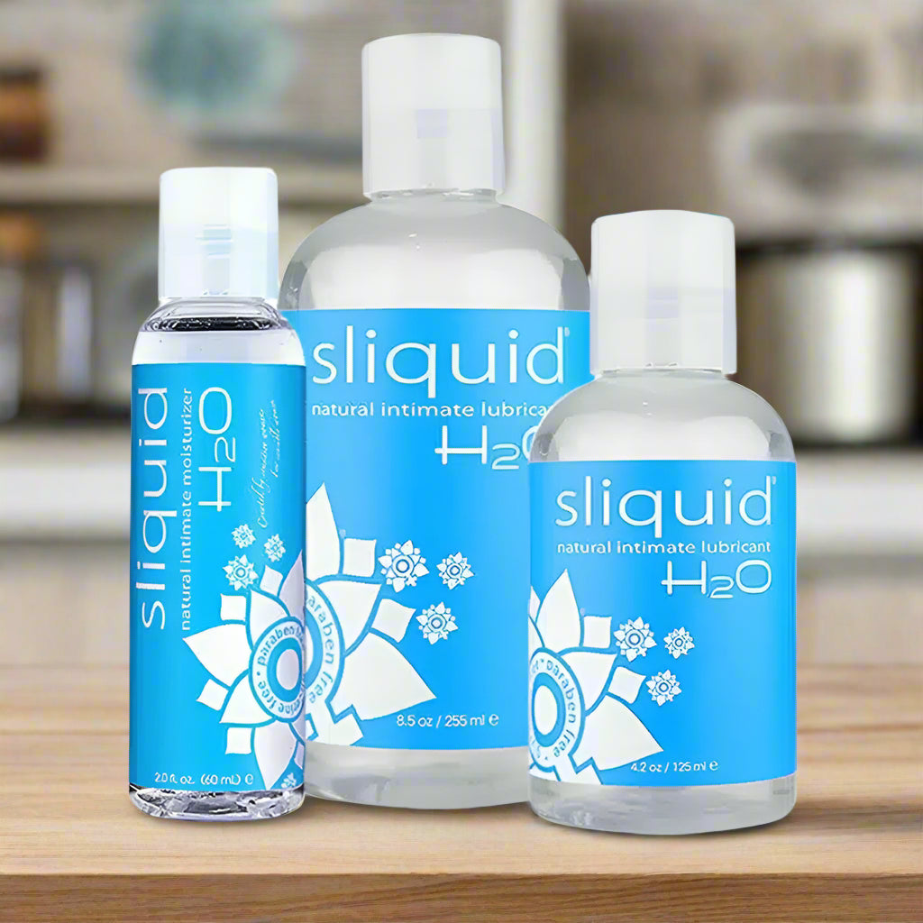 Three bottles of Sliquid H2O natural intimate lubricant of different sizes with blue and white labels are displayed on a kitchen counter with a blurred background.
