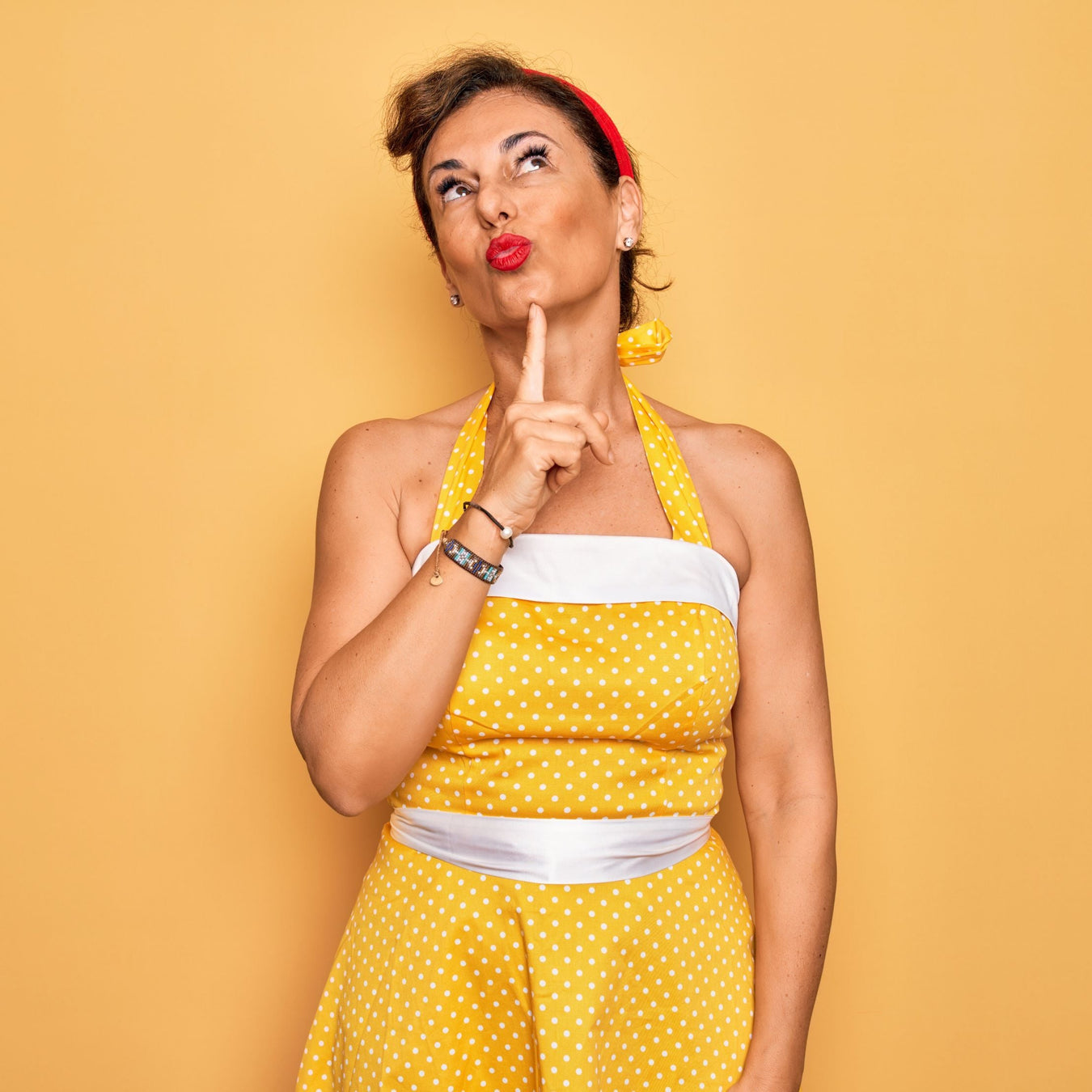 Woman in a yellow polka dot dress and red headband stands against a matching yellow background, looking up thoughtfully with her finger on her chin and lips pursed.