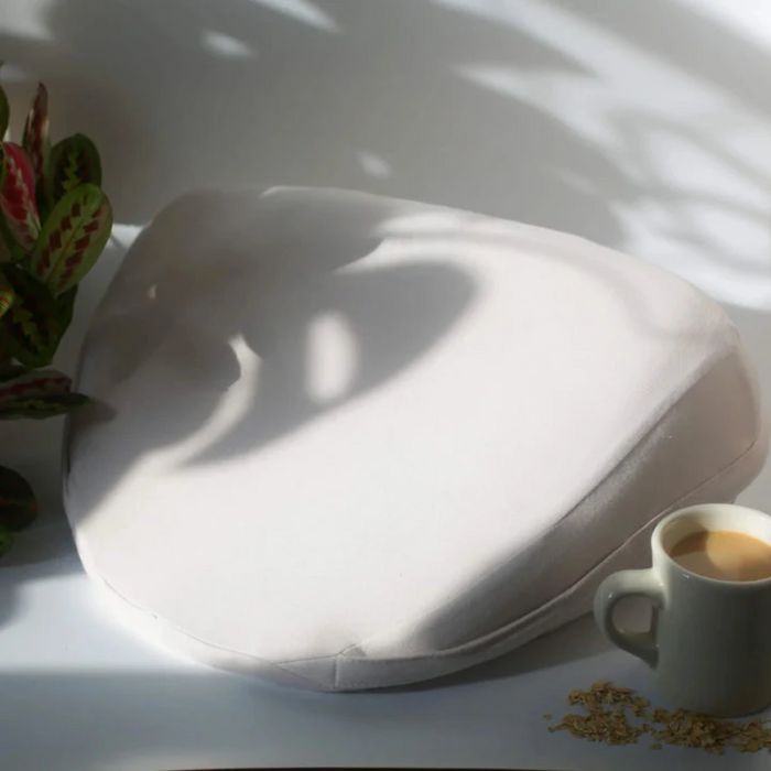 Off-white Dame Pillo positioning wedge pillow with a smooth, fabric cover, placed on a table next to a coffee mug.