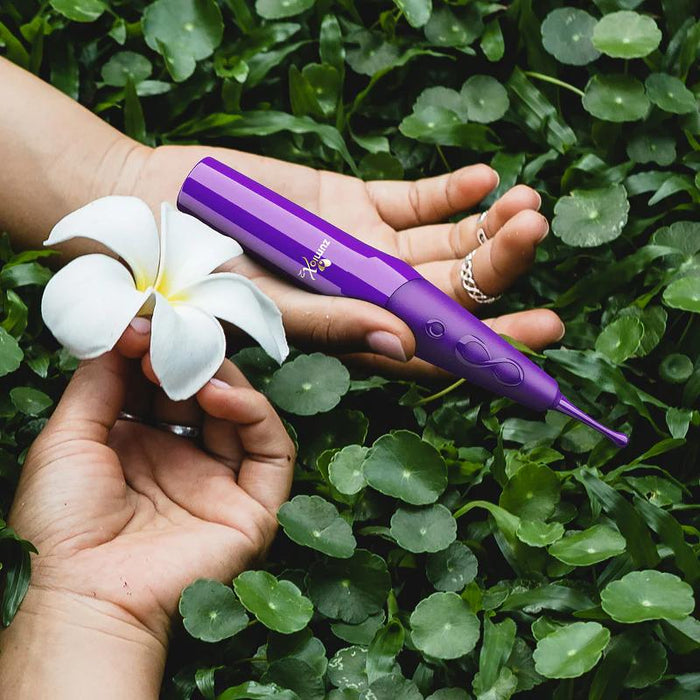 A person’s hands hold a white flower and the purple Zumio X2 Rotating Clitoral Stimulator against a backdrop of lush green leaves.