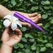 A person’s hands hold a white flower and the purple Zumio X2 Rotating Clitoral Stimulator against a backdrop of lush green leaves.