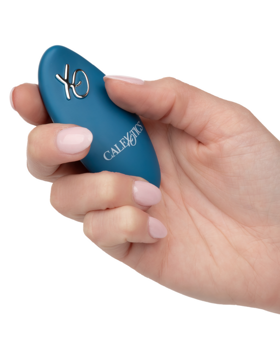 A hand with light pink manicured nails holds a small, blue, oval-shaped Dual Rider Remote Control Bump & Grind Humping Vibrator with the text "CalExotics" on its surface. The device has the letters "XO" in a metallic finish on one side. The background is white.