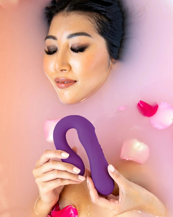 A woman relaxing in a bath with rose petals, holding a Jimmyjane Reflexx Rabbit 1 G-Spot & Clit Hugging Warming Vibrator - Purple from Pipedream Products, looking contented with closed eyes.