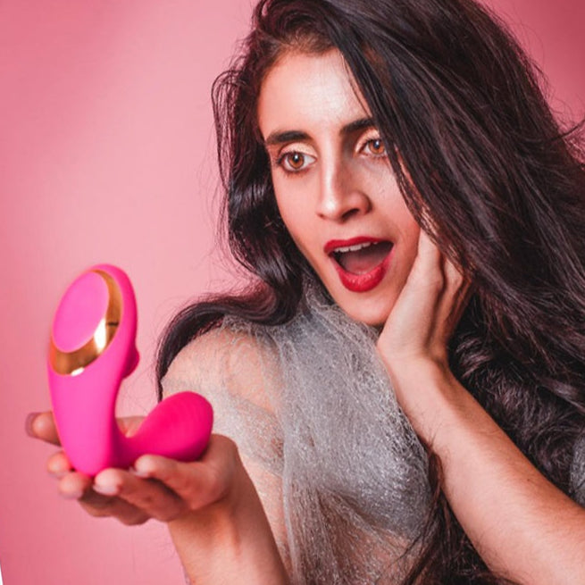 A woman with long dark hair and red lipstick looks surprised and intrigued as she holds Tracy's Dog OG Clitoral Sucking G-Spot Vibrator against a pink background.