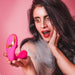 A woman with long dark hair and red lipstick looks surprised and intrigued as she holds Tracy's Dog OG Clitoral Sucking G-Spot Vibrator against a pink background.