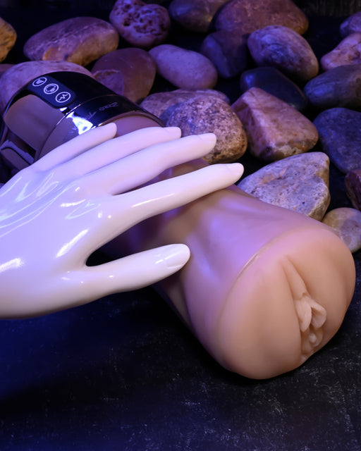 An XR Brands Tight Lipped Vibrating Vanilla Stroker with Suction + Free Movie Download with a flesh-toned exterior and a white robotic hand resting on it, set against a background of smooth round stones. The toy has a cylindrical shape with control buttons on its metallic top portion, offering varying vibrating speeds for enhanced pleasure.