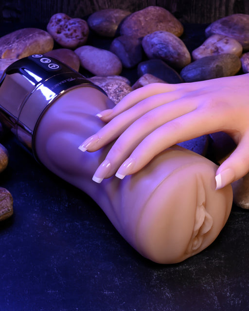 A hand with neatly manicured nails is resting on a flesh-colored, cylindrical object that resembles a sex toy. The Tight Lipped Vibrating Vanilla Stroker with Suction + Free Movie Download by XR Brands, placed on a dark surface with small stones in the background, features sucking functions and vibrating speeds. The scene is softly lit.