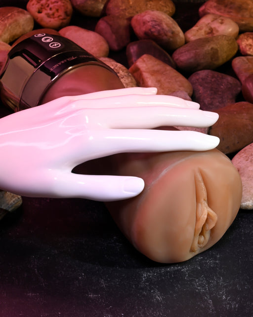 A white mannequin hand is placed on a flesh-colored Tight Lipped Vibrating Chocolate Stroker with Suction + Free Movie Download by XR Brands, designed to resemble female anatomy, set against a backdrop of various smooth stones. The detailed texture of the masturbator and the contrast with the hand are clearly visible.