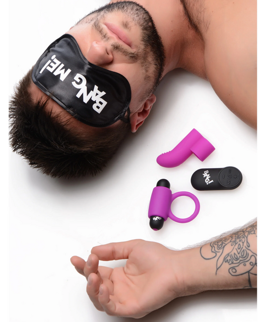 A person with short dark hair lies on a white surface, wearing the black blindfold from the BANG! Couple's Cock Ring, Finger Vibe, Bullet & Blindfold Kit - Purple by XR Brands that reads "BANG ME!". Next to them are three sex toys: a small purple vibrator, a purple ring vibrator, and a black remote-controlled bullet vibrator. The person's tattooed arm rests beside them.