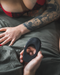 A close-up of a person holding a Hot Octopuss Atom Plus Dual Motor Vibrating Black Cock Ring while sitting on a bed, showcasing tattooed arms and wearing a red bra. The setting has a relaxed, intimate feel with soft bedding.