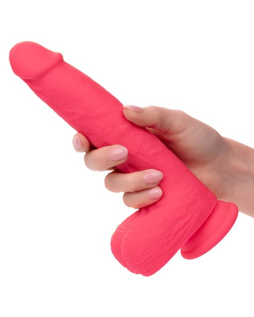 A person with light skin tone holds a CalExotics Silicone Stud Rumbling & Thrusting 9.5 Inch Pink Dildo with a suction cup base against a white background. The hand, adorned with neatly manicured nails, grips the silky silicone surface firmly.