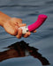 A hand holding a LELO Gigi 2 Silicone G-Spot Vibrator above the tranquil surface of water, casting a reflection.