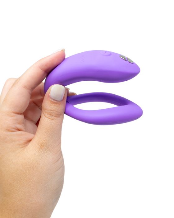 A hand holding a We-Vibe Sync O Hands-Free Wearable Couples Vibrator - Purple with a button and a looped structure, against a white background.