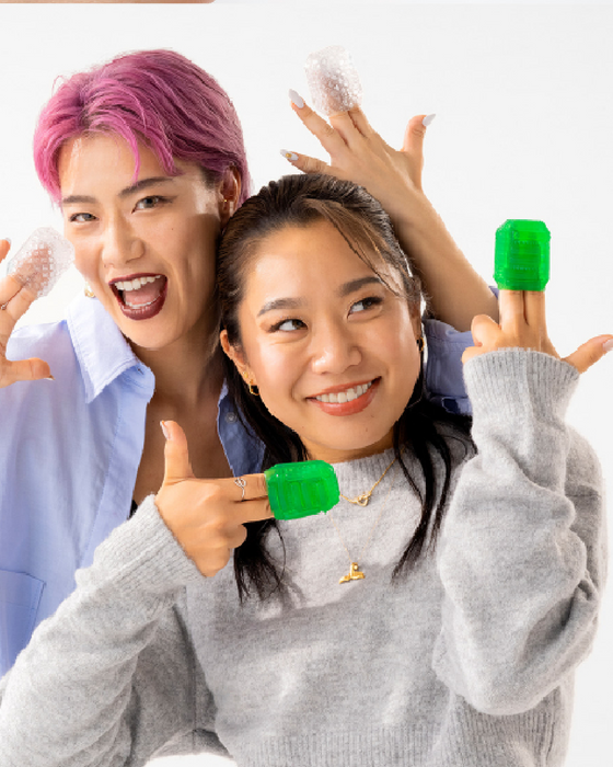 Two people smiling playfully at the camera, one with pink hair and the other with long black hair, both holding Tenga Uni Emerald Textured Finger Sleeves for Stroking and Clit Massage and green bottle caps. They appear joyful and engaged in a fun activity.