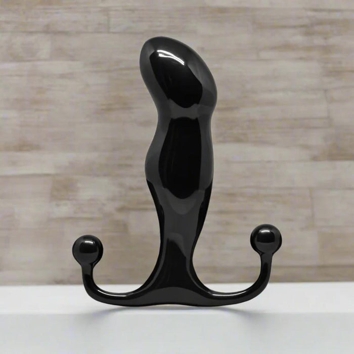 A shiny black, curved object with rounded ends stands upright on a white surface against a light, textured wall—this is the Aneros Progasm Black Ice Hands-Free Prostate Massager (Large), designed for hands-free stimulation.