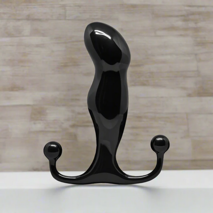 A shiny black, curved object with rounded ends stands upright on a white surface against a light, textured wall—this is the Aneros Progasm Black Ice Hands-Free Prostate Massager (Large), designed for hands-free stimulation.