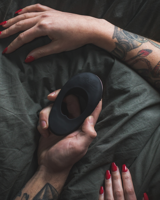 A close-up photo of two hands holding a Hot Octopuss Atom Plus Dual Motor Vibrating Black Cock Ring with an oval cutout, over a dark textured fabric. One hand has a tattoo and both have red-painted nails.