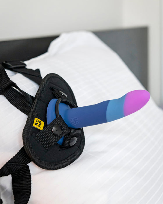 A prosthetic device with a Romp Piccolo First Time Pegging Kit - Strap on Harness + Dildo from Lovehoney, attached to a strap-on harness, resting on a white bedsheet.