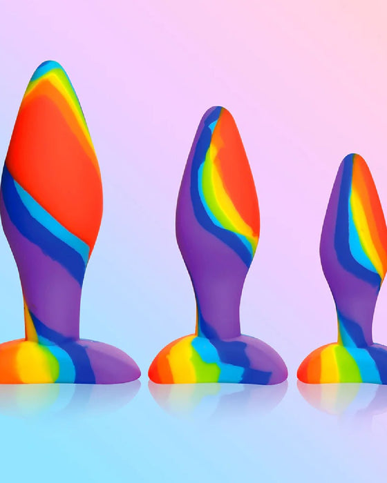 A trio of Simply Sweet Rainbow Silicone Butt Plugs by Curve Toys against a soft pink and blue gradient background.