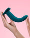 A person's hand holding a Fun Factory Limba Flex Medium Silicone Dildo in Deep Sea Blue with a suction base against a pink background. The device appears sleek and modern in design.