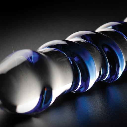 A close-up of the Icicles No. 5 Blue Spiral Glass 7 Inch Dildo shows its smooth, spiral borosilicate glass design resting on a dark surface, with reflections highlighting its glossy blue texture.
