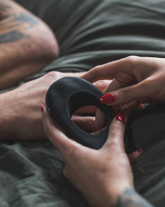 Close-up of a person with tattooed arms holding a Hot Octopuss Atom Plus Dual Motor Vibrating Black Cock Ring, set against a backdrop of gray bedding. The person has red-painted nails.
