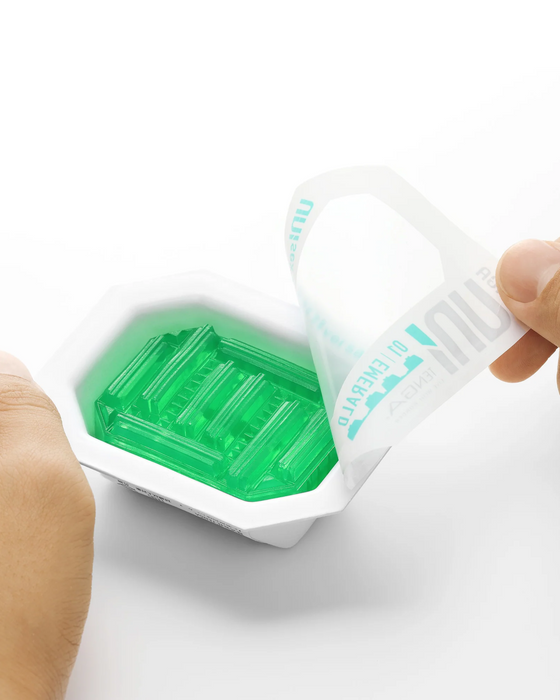 A person's hands peeling back the seal of a small white tray containing green gelatin. The seal has the words "Tenga Uni Emerald Textured Finger Sleeve for Stroking and Clit Massage" printed on it.