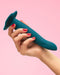 A hand with red nail polish holding a Fun Factory Limba Flex Medium Silicone Dildo - Deep Sea Blue against a pink background.