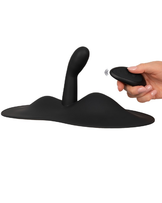 A hand holding a remote control operating an Orion VibePad 3 Ride On Hands-Free Humping Vibrator with G-Spot Probe, with a curved shape rising from a flat base.