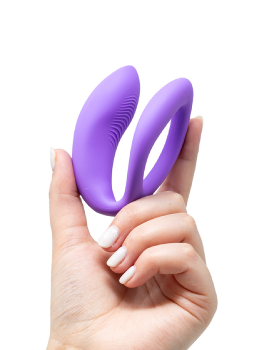 A hand holding a We-Vibe Sync O Hands-Free Wearable Couples Vibrator - Purple with a dual loop design against a white background.
