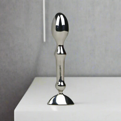 The Aneros Tempo Stainless Steel Anal Stimulator stands upright on a white surface against a plain gray background, showcasing its metallic, reflective finish and ergonomic design—ideal for temperature play.
