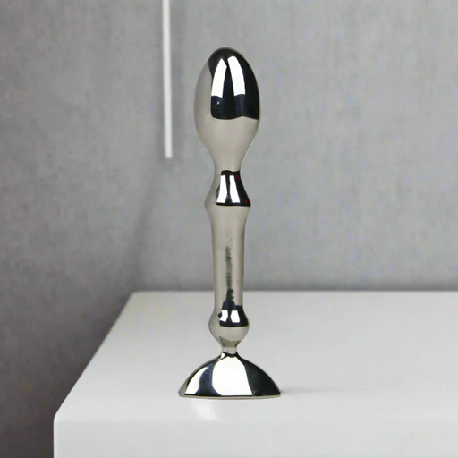 The Aneros Tempo Stainless Steel Anal Stimulator stands upright on a white surface against a plain gray background, showcasing its metallic, reflective finish and ergonomic design—ideal for temperature play.