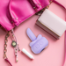 A flat lay on a pink background features a pink purse, light pink wallet, charm-strap wristwatch, tube of lip balm, and the M'Lady Rotating and Licking Tongue Vibrator in lavender.
