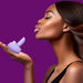 A woman with smooth brown skin and straight hair holds the M'Lady Rotating and Licking Tongue Vibrator in her palm, puckering her lips against a solid purple background.