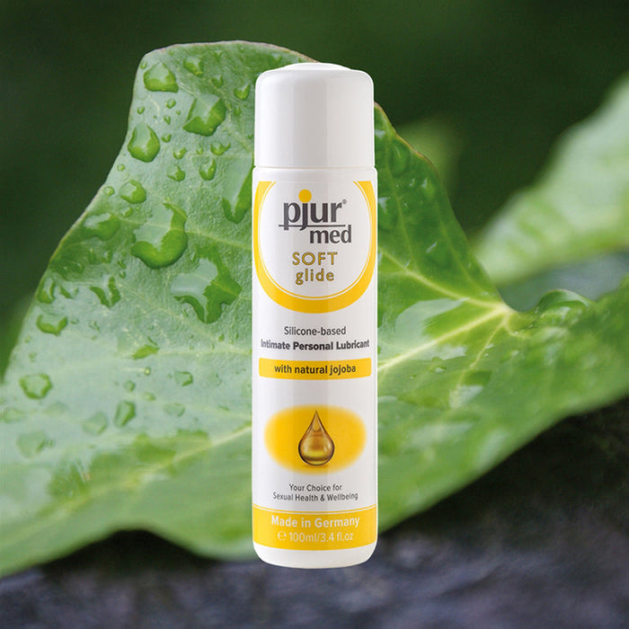 A white bottle of Pjur Med Soft Glide Silicone Lubricant with Jojoba Oil stands upright on a water-dappled green leaf. Designed for sensitive intimate areas, the bottle has yellow, gray, and white labeling.