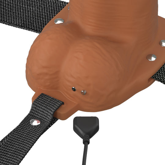 A close-up of the Fantasy X-Tensions Elite Vibrating Hollow 6 Inch Silicone Dildo & Strap-On Harness in Chocolate, showing its silicone shaft, adjustable straps, and a small charging port being connected to a black charger.