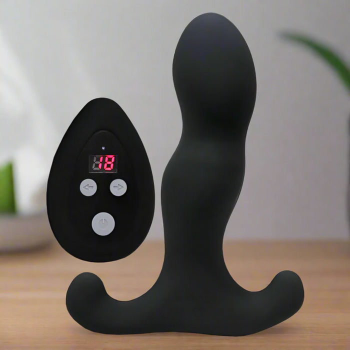 The Aneros Vice 2 Vibrating Remote Control Prostate Massager, in black with a curved design, rests on a wooden surface beside its black remote showing “18” on the LED screen. The softly blurred background features green and white tones.
