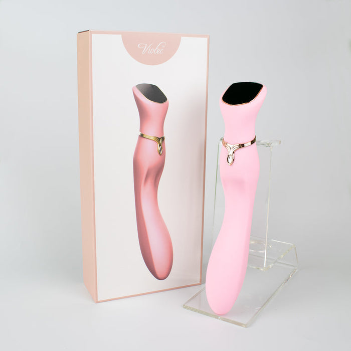 The Chance Waterproof Touch Screen G-Spot Vibrator, made from medical-grade silicone, is pink with a gold accent and stands upright on a clear stand beside its matching box featuring the product image against a light background.