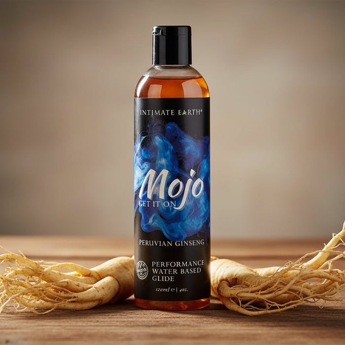 A bottle of Mojo Performance Water-Based Glide With Peruvian Ginseng sits on a wooden surface, flanked by two pieces of ginseng root.
