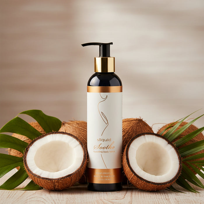 A bottle of Sliquid Soothe Sweet Coconut Hydrating Body Lotion with a gold pump sits between two halved coconuts and green palm leaves on a light wooden surface, against a softly blurred wooden background.