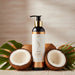 A bottle of Sliquid Soothe Sweet Coconut Hydrating Body Lotion with a gold pump sits between two halved coconuts and green palm leaves on a light wooden surface, against a softly blurred wooden background.