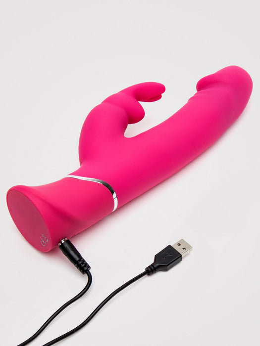 The Happy Rabbit Dual Density Rechargeable Silicone Rabbit Vibrator, featuring dual stimulation arms in pink, rests on a white surface with its USB charging cable attached.