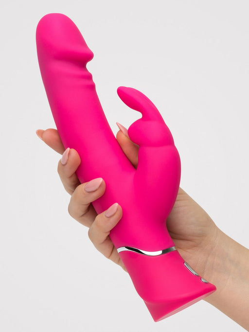 A hand holds the Happy Rabbit Dual Density Rechargeable Silicone Rabbit Vibrator, a bright pink lifelike silicone toy with a curved design and smaller external stimulator, against a plain light background.