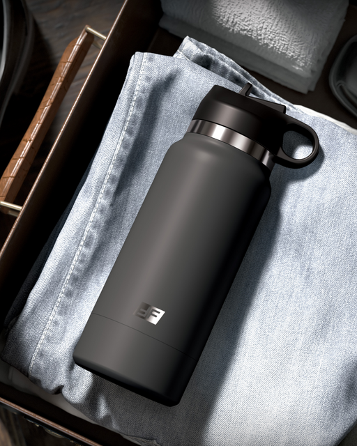 A black Fuck Flask Pussy Stroker Disguised as a Water Bottle - Vanilla by Pipedream Products with a loop cap is placed on top of a pair of folded light blue jeans in a wooden drawer. Nearby are neatly arranged towels and other items, all meticulously organized. The water bottle, bearing an "EE" logo on its side, remains discreet within the tidy setting.