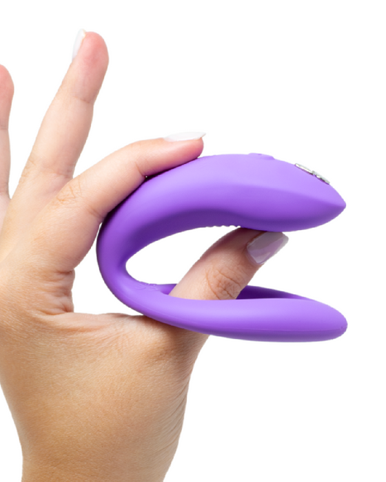 A hand holding a We-Vibe Sync O Hands-Free Wearable Couples Vibrator in purple silicone against a white background.